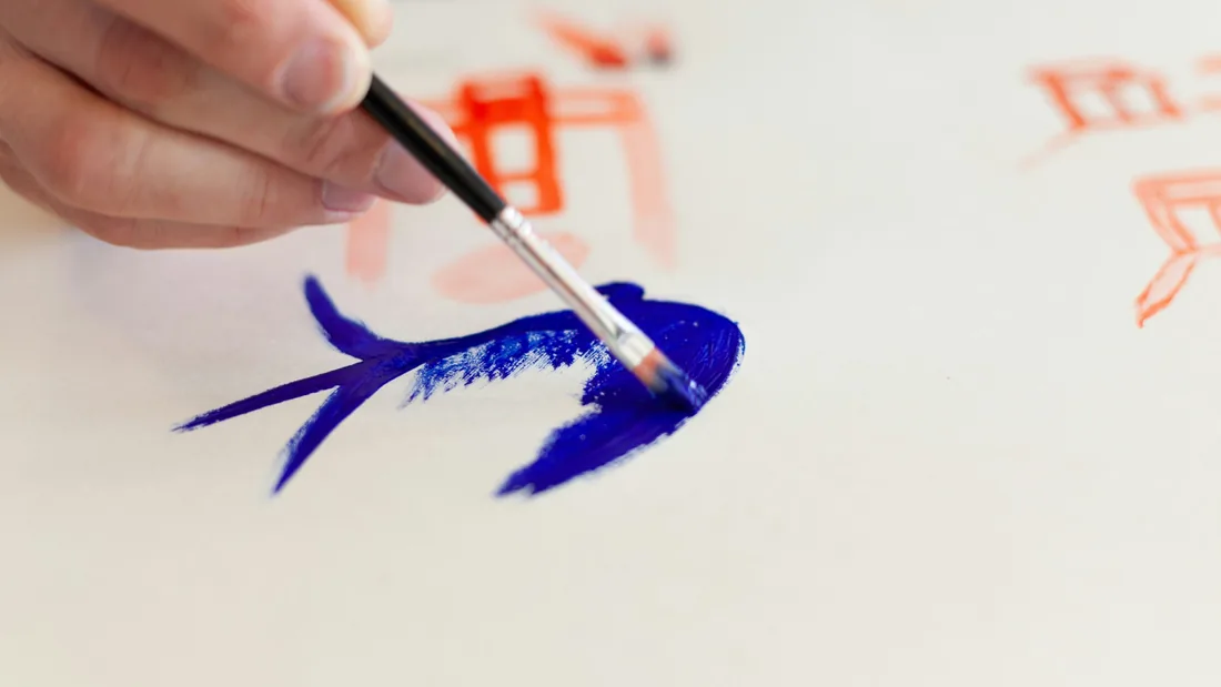 Mano sosteniendo un pincel fino que traza una letra con pintura azul sobre una superficie clara, junto a un boceto de letras en rojo